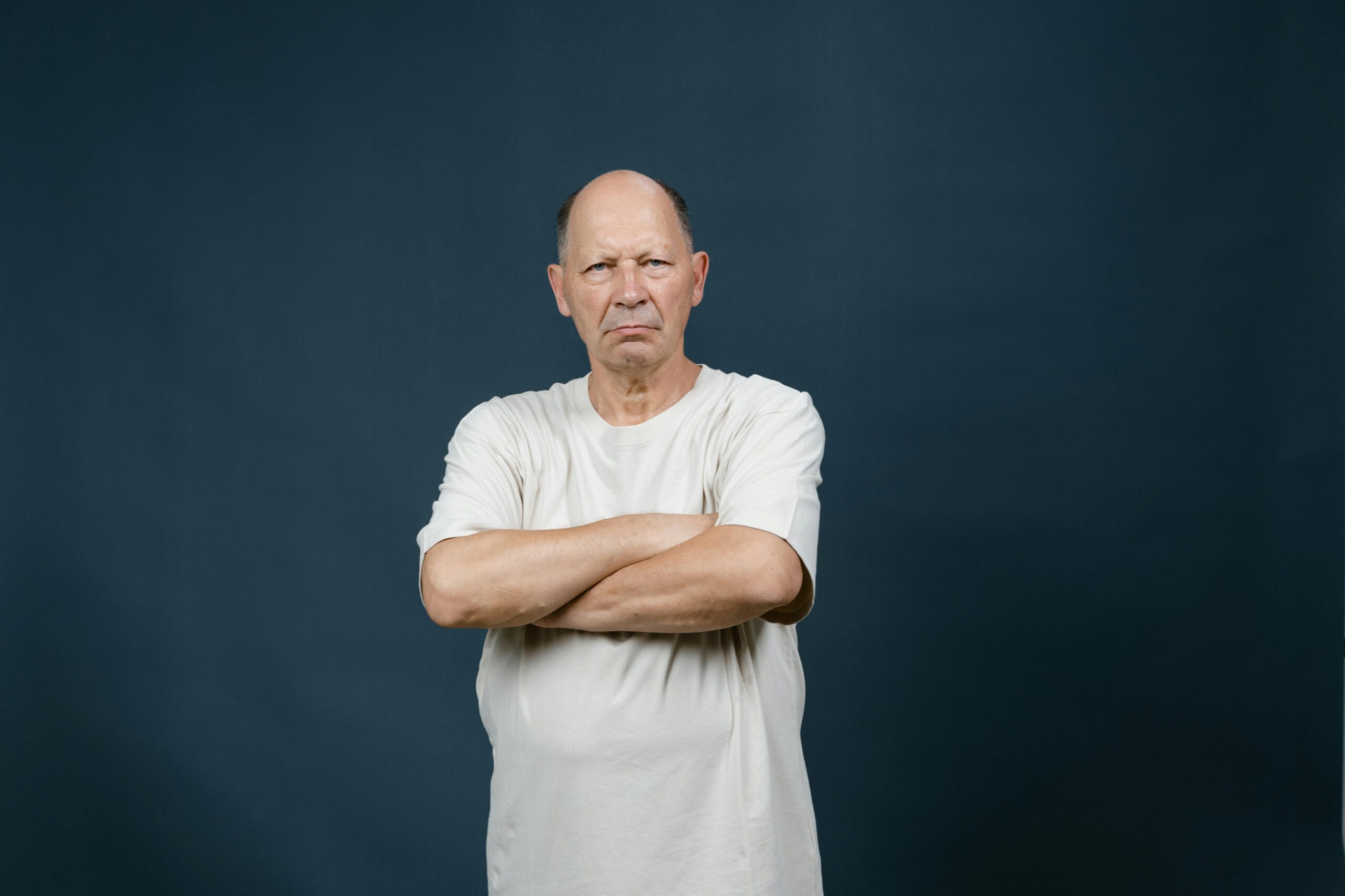 A senior man in a white shirt with a serious expression crossing arms against a dark background.